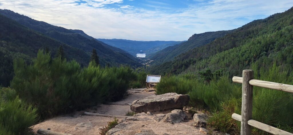A rocky viewpoint with a wooden fence and an information board, looking down a forested valley to a distant lake.