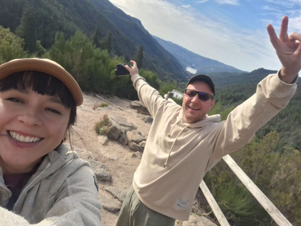 Two people taking a happy selfie with arms raised at the viewpoint, hills and a lake behind them.