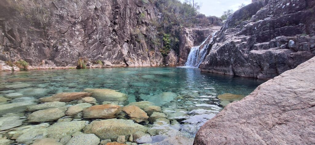 A small waterfall falling between dark granite walls into a clear turquoise pool with smooth rounded boulders.