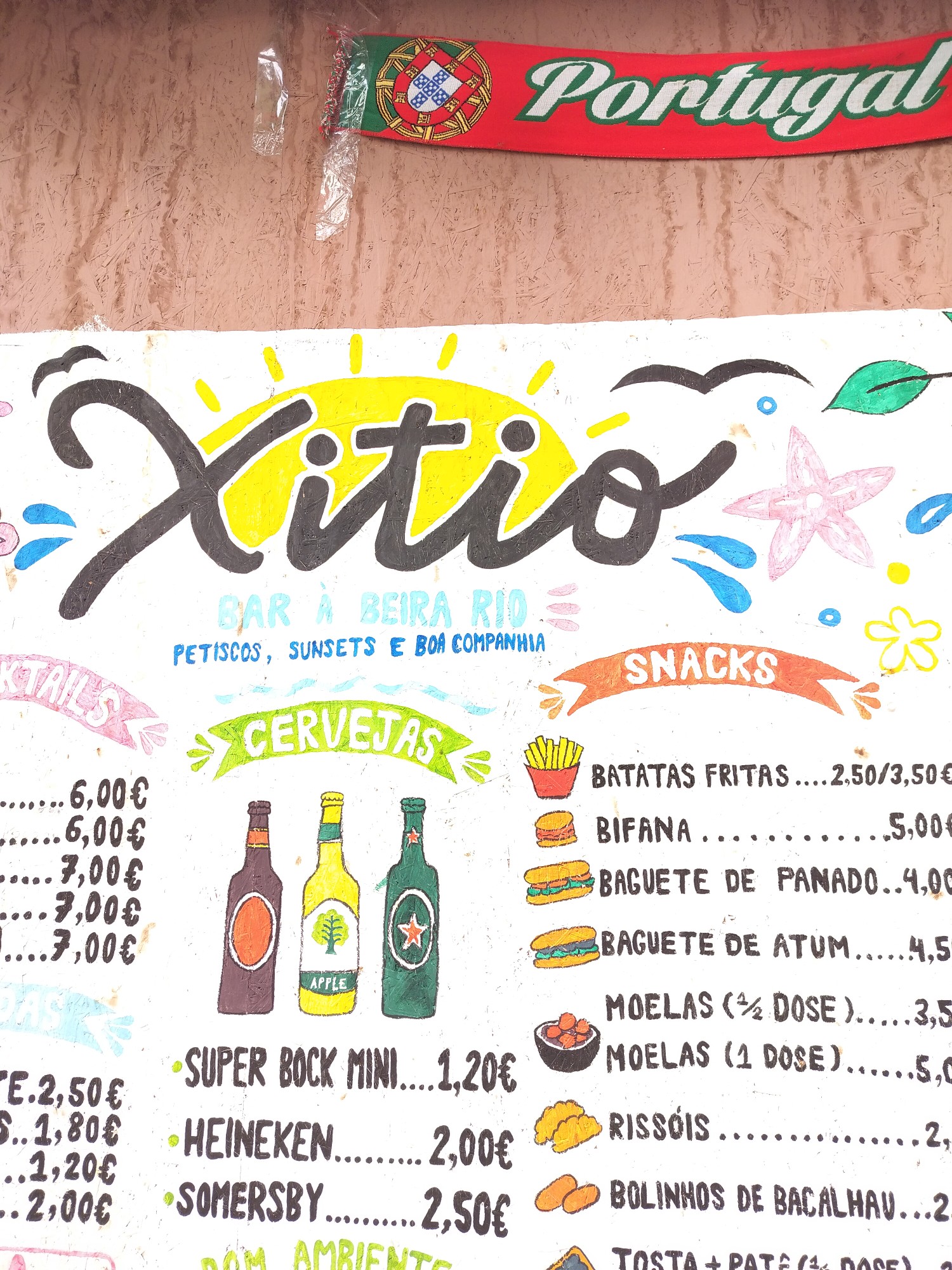 Hand-painted menu board of the bar Xitio: the name in black script over a yellow sun, with the words "bar à beira rio, petiscos, sunsets e boa companhia" beneath.