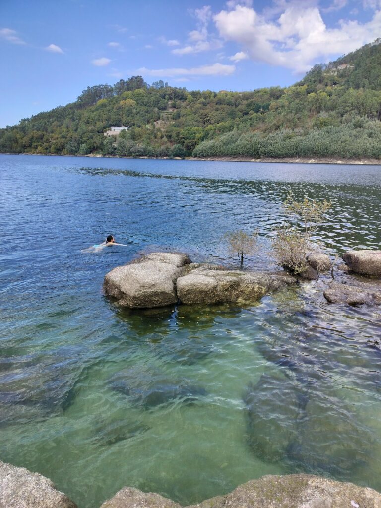 Clear green water of the reservoir with a rocky islet, a swimmer, and forested hills under a blue sky with white clouds.